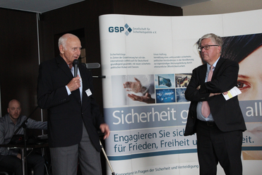 Zwei Männer auf einer Veranstaltung der Gesellschaft für Sicherheitspolitik (GSP); einer spricht ins Mikrofon, der andere hört zu.