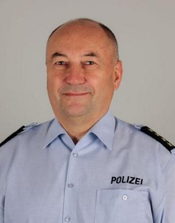 Kopfaufnahme eines lächelnden, mittelalten Mannes in einer deutschen Polizeiuniform mit 'POLIZEI' auf der Tasche und Schulterklappen.