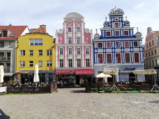 Farbige historische Gebäude in Gelb, Rosa und Blau mit Ziergiebeln säumen einen gepflasterten Platz mit Straßencafés.