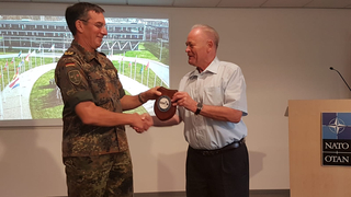 Ein deutscher Soldat in Uniform überreicht einem lächelnden Mann eine GSP-Plakette und schüttelt seine Hand bei einem NATO-Event.