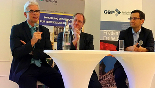 Drei Männer in Anzügen bei einer Podiumsdiskussion vor Bannern zu Forschung, Verteidigung und Sicherheit.