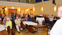 Eine Seniorengruppe hört einem Mann zu, der in einem Versammlungssaal in ein Mikrofon spricht, mit Publikum und Projektor.