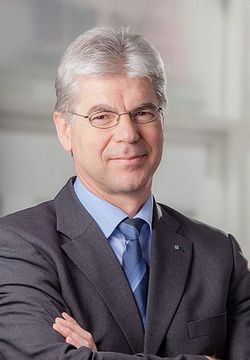 Porträt eines lächelnden Mannes mit grauem Haar und Brille, der einen dunklen Anzug und eine blaue Krawatte trägt, Arme verschränkt.