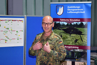 Lächelnder Bundeswehr-Offizier Merz in Tarnuniform vor Tafeln zu taktischer Simulation und Luftbeweglichkeitstraining.