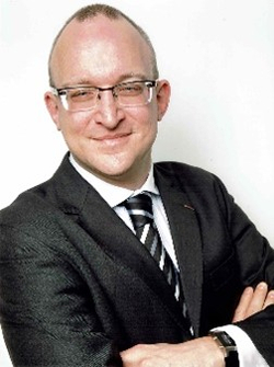 Ein lächelnder Mann in dunklem Anzug mit Brille und verschränkten Armen.