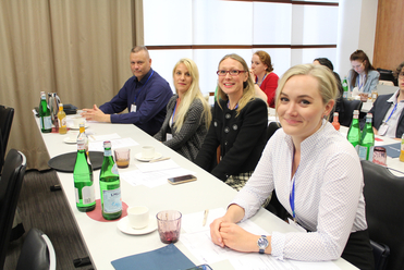 Teilnehmer an einem Konferenztisch mit Erfrischungen. Eine lächelnde blonde Frau im Vordergrund blickt in die Kamera.