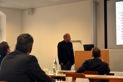 Ein Mann präsentiert einem Publikum in einem Seminarraum, mit Teilnehmern von hinten an Tischen sichtbar.