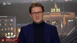 Nachrichtensprecher mit Brille berichtet über russischen Angriff auf die Ukraine, mit Brandenburger Tor im Hintergrund.