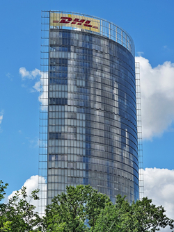 Der hohe, gekrümmte Glas-DHL-Wolkenkratzer vor blauem Himmel mit Wolken, teilweise von Bäumen verdeckt.