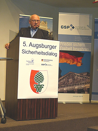 Mann spricht am Rednerpult beim 5. Augsburger Sicherheitsdialog, mit Bannern, die das Augsburger Wappen und deutsche Symbole zeigen.