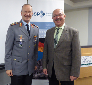 Zwei lächelnde Männer, einer in Militäruniform, der andere im Anzug, vor einem GSP-Banner und Rednerpult.