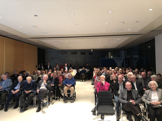 Ein vielfältiges Publikum, hauptsächlich Erwachsene und Senioren, sitzt in einem modernen Konferenzraum bei einer Präsentation.