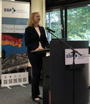 Lächelnde Frau mit Brille spricht an einem Rednerpult. Dahinter ein GSP-Banner mit Deutschlandfahne und Reichstagsgebäude.