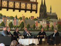 Podiumsdiskussion mit sechs Männern vor einem Wandgemälde der Kölner Altstadt und des Doms.