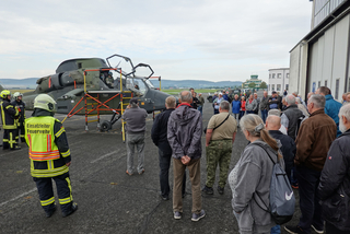 Menschengruppe besichtigt einen Eurocopter Tiger Kampfhubschrauber auf einem Flugfeld, mit Feuerwehrleuten im Vordergrund.