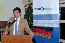 Mann spricht am Rednerpult vor einem Banner der Gesellschaft für Sicherheitspolitik (GSP) mit deutscher Flagge.