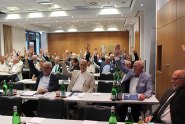 Gruppe von hauptsächlich Männern in einem Konferenzraum, die ihre Hände zum Abstimmen erheben.