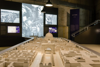 Modell einer monumentalen Stadtplanung in einem Museum. Im Hintergrund zeigen Bildschirme historische Aufnahmen von Deportation und Zwangsarbeit.