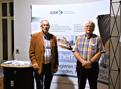 Zwei Männer stehen vor einem Banner der "Gesellschaft für Sicherheitspolitik e.V." bei einer Veranstaltung.