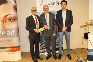 Drei Männer posieren bei einer GSP-Veranstaltung. Der Mann links hält ein Geschenk, im Hintergrund GSP-Banner.