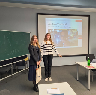 Zwei junge Frauen in einem Seminarraum. Links eine Tafel mit 'UN Security Council', rechts ein Bildschirm mit 'Questions? Feedback?'.