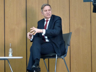 Ein Mann im dunklen Anzug, weißem Hemd und roter Krawatte sitzt gestikulierend und sprechend bei einer Veranstaltung.