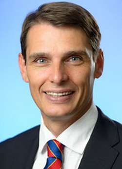 Porträt eines lächelnden Mannes mittleren Alters mit kurzen braunen Haaren, blauen Augen, Anzug und gestreifter Krawatte.