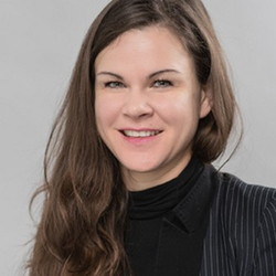 Professionelles Porträt einer lächelnden Frau mit langen braunen Haaren, gestreiftem Blazer und schwarzem Oberteil vor grauem Hintergrund.