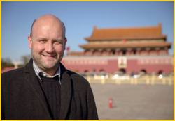 Lächelnder Glatzkopf mit Bart in schwarzem Mantel vor dem verschwommenen Tor des Himmlischen Friedens in Peking.