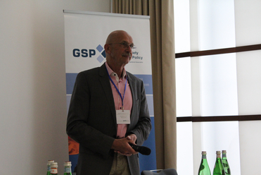 Glatzköpfiger Mann mit Brille spricht in Mikrofon bei einer GSP-Konferenz, mit Markenbanner im Hintergrund.