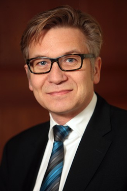 Porträt eines lächelnden Mannes mit hellerem Haar, Brille, dunklem Anzug und blau-grau gestreifter Krawatte.