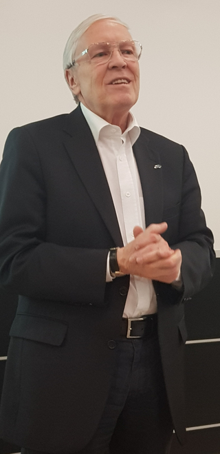 Ein älterer Mann mit weißem Haar und Brille lächelt und hält die Hände gefaltet, trägt einen dunklen Anzug und weißes Hemd.