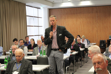 Ein Mann spricht mit Mikrofon vor einem sitzenden Publikum in einem Konferenzraum. Wasserflaschen stehen auf Tischen bereit.