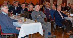 Eine Gruppe Männer bei einer Veranstaltung, an Tischen in einem Saal sitzend, einige mit Getränken, nach vorne blickend.