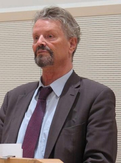 Mann mit grauem Haar und Bart in dunklem Anzug und Krawatte, blickt nachdenklich nach links.
