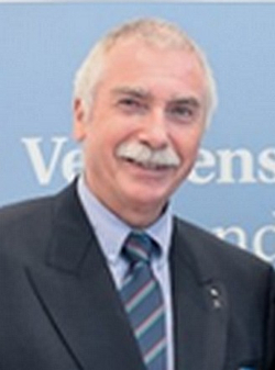 Lächelnder Mann mit weißen Haaren und markantem Schnurrbart, Anzug und gestreifter Krawatte, vor hellblauem Hintergrund.