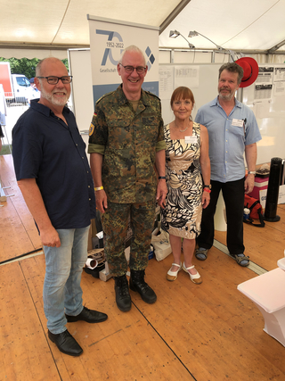 Vier lächelnde Personen: zwei Männer (einer in Militäruniform) und eine Frau, posieren auf einer Veranstaltung mit einem Banner.