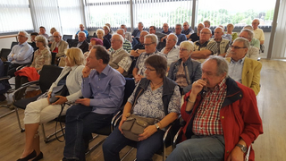 Eine Gruppe meist älterer Erwachsener sitzt in Reihen in einem hellen Raum und blickt nach vorne, während sie einer Veranstaltung lauschen.