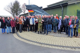 Große Gruppe Männer, darunter ein Soldat, posiert draußen vor einem deutschen Flugzeug-Leitwerk. Zwei Männer schütteln Hände.