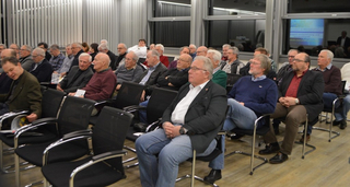 Zahlreiche ältere Männer sitzen in einem Raum und lauschen einem Vortrag oder einer Präsentation.