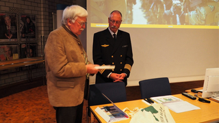 Ein älterer Mann im Trachten-Sakko überreicht Papiere an einen Marineoffizier bei einer Präsentation über Militär.