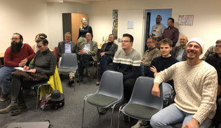 Vielfältige Gruppe von Menschen, hauptsächlich Männer, nimmt an einem Treffen oder einer Präsentation in einem hellen Raum teil, einige lächeln.