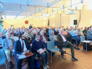 Ein großes Publikum aus Erwachsenen und Senioren sitzt in Reihen und hört aufmerksam in einem hellen Konferenzraum zu.