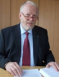 Ein älterer Mann mit weißem Bart und Brille, gekleidet in Anzug und roter Krawatte, sitzt lächelnd an einem Schreibtisch mit Dokumenten.
