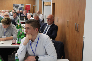 Nahaufnahme eines jungen Mannes, der auf einer Konferenz aufmerksam zuhört, mit anderen Teilnehmern und Tischen mit Getränken im Hintergrund.