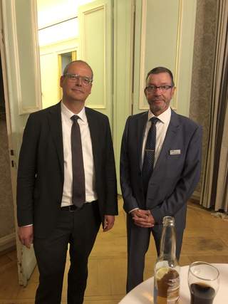 Zwei Männer in Anzügen in einem eleganten Raum. Einer mit dunklem, der andere mit blauem Anzug und Namensschild. Getränke im Vordergrund.