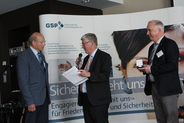 Drei Männer bei einer Veranstaltung der Gesellschaft für Sicherheitspolitik. Ein Mann präsentiert eine Auszeichnung.