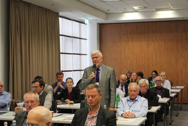 Ein Mann mit grauem Bart spricht bei einer Konferenz ins Mikrofon und wendet sich an ein sitzendes Publikum.