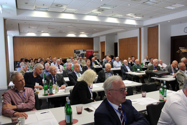 Teilnehmer, meist ältere Männer, sitzen an Tischen in einem hellen Konferenzraum während einer Veranstaltung.
