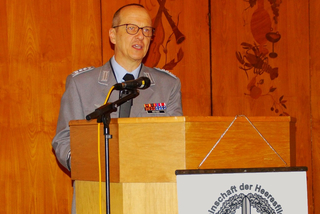 Deutscher Offizier RÜTHER in Uniform und Brille spricht an einem Rednerpult mit Mikrofon vor einer dekorierten Holzwand.